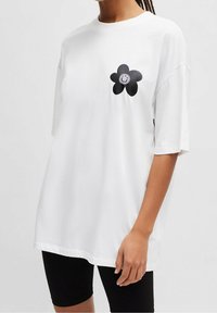 Biała, oversize'owa koszulka z bawełny, z czarnym kwiatem z uśmiechniętą twarzą na piersi. Krótkie rękawy i luźny krój.