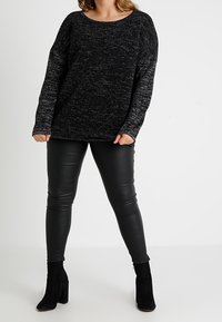 Žena v černém volném svetr s melírovaným vzorem, černých legínách s koženým vzhledem a černých semišových kotníkových botách stojí na bílém pozadí.