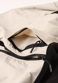 Tasca con cerniera su esterno in tessuto beige con accenti in pile nero. Include cerniere nere a contrasto e linguette di estrazione, che mostrano un design funzionale.