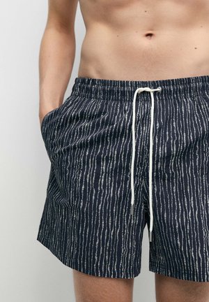 Personne torse nu portant un short de bain noir à fines rayures verticales blanches et cordon blanc, une main dans la poche, vue de la taille à la cuisse.