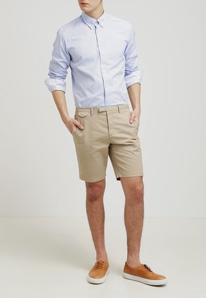 Homme portant une chemise à manches longues bleu clair retroussées, un short beige et des baskets beige, debout les mains dans les poches.