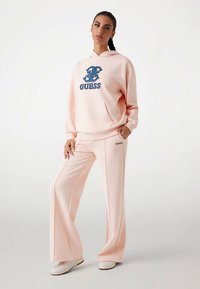 Sudadera rosa con logo azul "GUESS", combinada con pantalones anchos rosa a juego. El material parece suave. Modelo en postura casual.