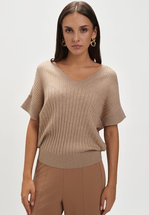 Beżowy sweter z prążkowanego materiału w serek, z krótkimi rękawami, z dopasowanym dolnym brzegiem i miękką teksturą, zestawiony z spodniami w podobnym odcieniu.