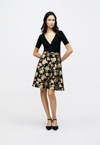 Μαύρο φόρεμα τύπου wrap με κοντά μανίκια, διαθέτει φουσκωτή φούστα διακοσμημένη με χρυσά λουλουδένια σχέδια. Λείο ύφασμα με V-λαιμόκοψη.