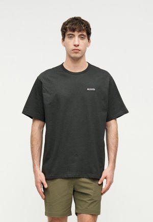 Junger Mann, der nach vorne blickt, trägt ein schwarzes Columbia-T-Shirt und olivgrüne Shorts vor einem schlichten weißen Hintergrund.