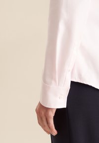 Polsino di camicia in cotone rosa chiaro con una superficie liscia e un motivo a tessitura diagonale. Presenta un singolo bottone sul polsino. Tessuto scuro sottostante.