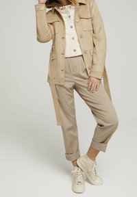 Giacca in suede beige con tasche frontali e chiusura a bottone, abbinata a pantaloni beige chiaro. Sneakers bianche completano l'outfit.