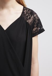 Blusa nera con maniche corte in pizzo, scollatura a V, tessuto morbido, parte anteriore plissettata e delicati dettagli in pizzo sulle cuciture delle spalle.