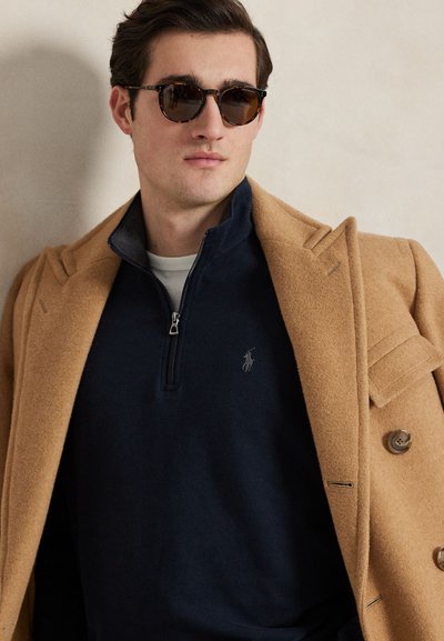 Homme portant des lunettes de soleil en écaille, un pull bleu marine à zip court avec un logo, et un manteau croisé camel posé sur les épaules devant un mur neutre.