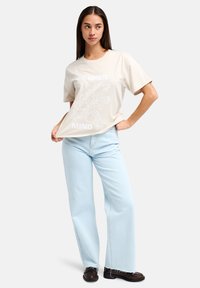 Μπεζ βαμβακερό μπλουζάκι με λευκό γράφημα "OPEN MIND" και αφηρημένα σχέδια. Συνδυασμένο με ανοιχτόχρωμα μπλε τζιν παντελόνια με πλατύ πόδι και μαύρα παπούτσια.