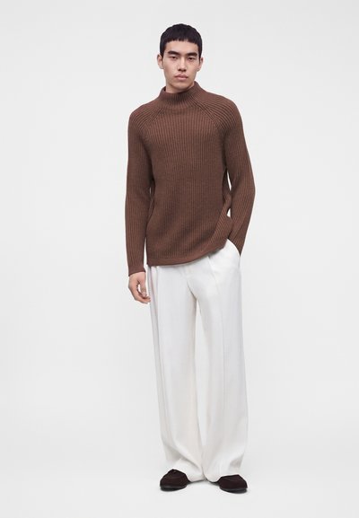 Jeune homme debout portant un pull à col roulé côtelé marron, un pantalon large blanc et des chaussures foncées sur un fond clair uni.