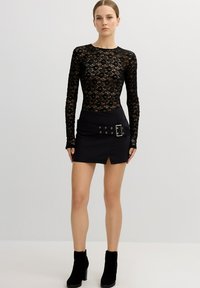 Bodysuit de encaje negro con mangas largas, combinado con una falda mini negra que presenta una hebilla decorativa y detalle de abertura, botines negros con tacón.