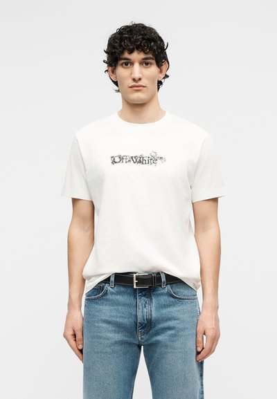 Νεαρός άνδρας με σγουρά μαλλιά που φοράει λευκό t-shirt Off-White, μπλε τζιν και μαύρη ζώνη, στέκεται μπροστά σε απλό φόντο.