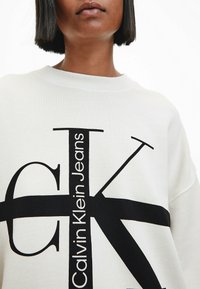 Sudadera blanca con un diseño gráfico negro que presenta las letras "CK" y "Calvin Klein Jeans". Cuello acanalado y textura suave.