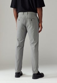 Pantalon gris en tissu texturé, jambes fuselées, et deux poches arrière avec fermetures à bouton. Porté avec des chaussures noires.