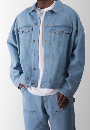Mand iført lys blå denimjakke og matchende jeans over en hvid t-shirt, med den ene hånd i lommen og den anden hvilende ved siden.