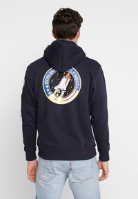 Sweatshirt cu glugă de culoare bleumarin, fabricat din bumbac, având un logo cu o navetă spațială pe spate, cu manșete și tiv deshise.