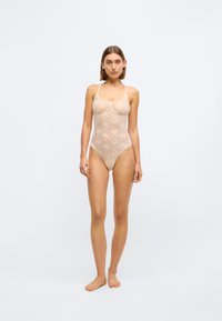 Spitzen-Bodysuit mit floralem Muster in sanftem Beige, ausgestattet mit verstellbaren Trägern und transparenter Textur, entworfen mit einem tiefen Ausschnitt.