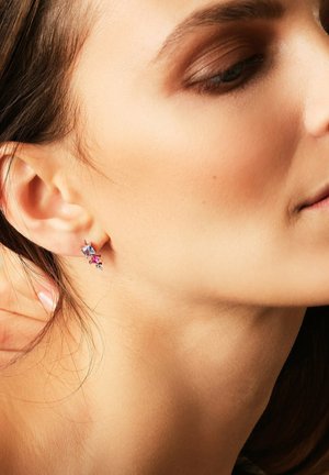 Ohren einer Frau mit einem silbernen Ohrring, der blaue, rosa und klare Edelsteine zeigt, teilweise sichtbares Gesicht, das leicht zur Seite gedreht ist.