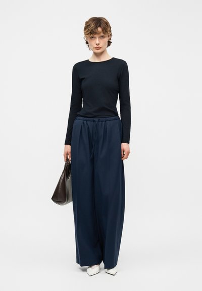 Γυναίκα που φοράει μαύρο τοπ με μακριά μανίκια, σκούρο μπλε φαρδιά παντελόνια, λευκά μυτερά παπούτσια, κρατώντας καφέ σκούρο τσάντα στο χέρι, στέκεται μπροστά σε λευκό φόντο.