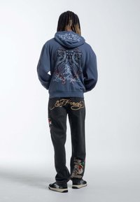 Blue hoodie with graphic design featuring two serpents and text, paired with black jeans adorned with colorful embroidered skull and floral patterns.