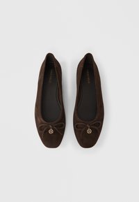 Καφέ μπαλαρίνες από σουέντ με στρογγυλεμένη μύτη, διακοσμημένες με φιόγκο και χρυσό διακοσμητικό. Λείο εσωτερικό επένδυμα και επίπεδη σόλα.