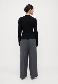 Vrouw met een donkere gevlochten bun, gekleed in een zwarte blouse met lange mouwen en grijze wijde broek met strepen, staand met de rug naar de camera tegen een neutrale achtergrond.