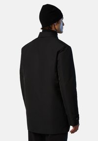Black waterproof coat with a high collar, smooth texture, buttoned cuffs, and a straight silhouette, worn with a matching black beanie.