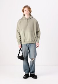 Light green hoodie with a front pocket, paired with distressed blue jeans and chunky black shoes. Model holds a black shoulder bag.