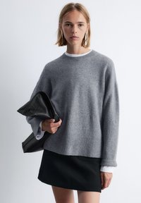 Grauer Strickpullover mit geripptem Saum und Bündchen, kombiniert mit einem schwarzen Rock. Das Model hält eine strukturierte schwarze Clutch. Weicher, lässiger Schnitt.