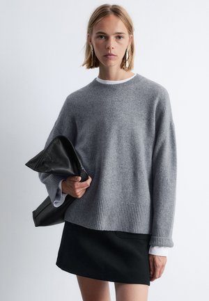 Gray knitted sweater with ribbed hem and cuffs, paired with a black skirt. Model holds a textured black clutch. Soft, relaxed fit.