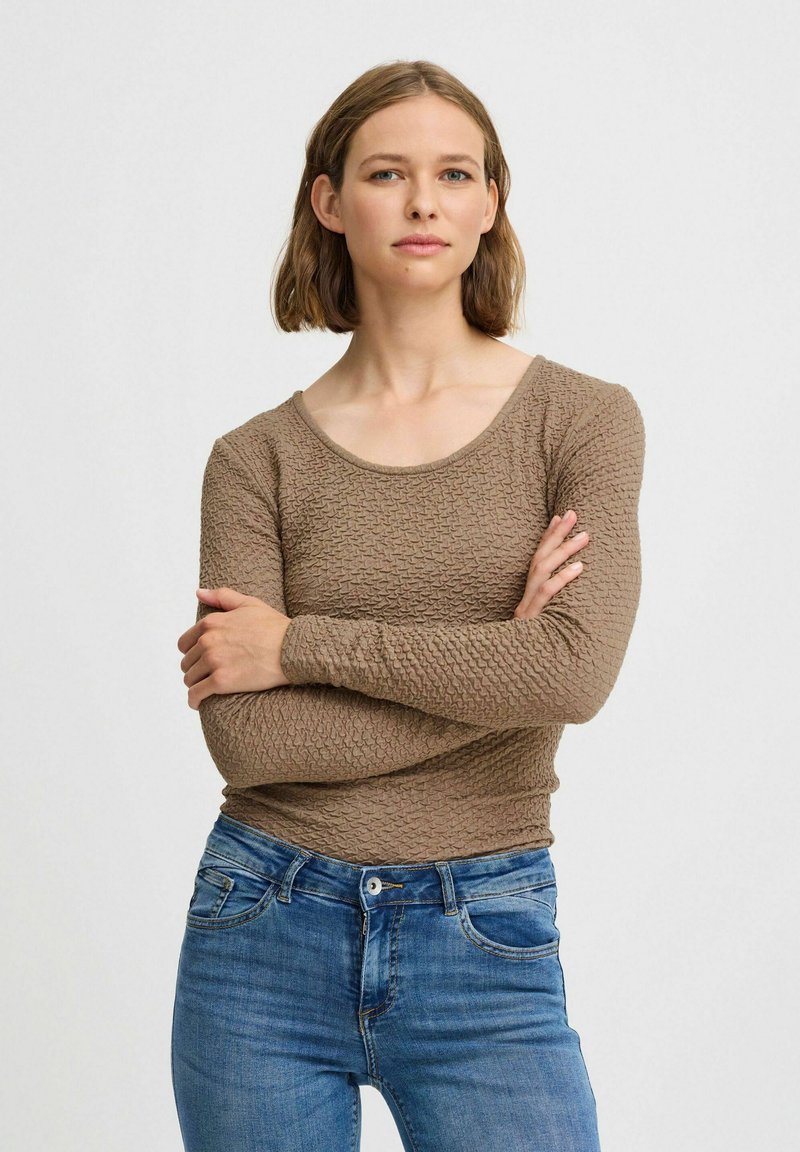 Långärmad, ljusbrun topp i strukturerad tyg, rund halsringning och figurnära design, kombinerad med blå jeans.