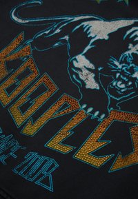 Black fabric with a roaring panther illustration in white and blue, above glittery gold letters spelling "KENZO" and blue text below.