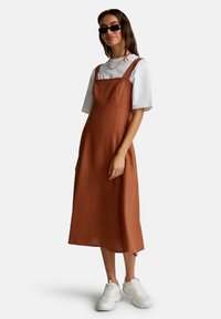 Mujer vistiendo un vestido midi sin mangas color óxido sobre una camiseta blanca, zapatillas blancas con suela gruesa, gafas de sol negras y collar de cadena dorada.