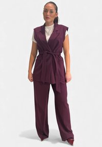 Gilet sartoriale color marrone scuro con cintura annodata sopra una maglia bianca, abbinato a pantaloni a gamba larga coordinati. Tessuto liscio con design strutturato.