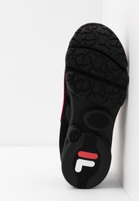 Semelle de chaussure de sport noire avec une adhérence en caoutchouc texturé, présentant des accents rouges et un logo en blanc et rouge. Le côté a une finition lisse noire.