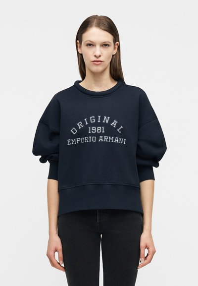 Femme aux cheveux raides portant un sweat-shirt bleu marine avec le texte « ORIGINAL 1981 EMPORIO ARMANI » et un pantalon noir, debout devant un fond blanc.