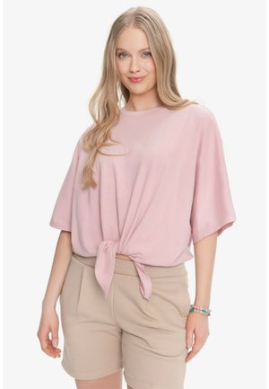 Młoda kobieta z długimi blond włosami, ubrana w luźną różową związaną koszulę i beżowe szorty, stoi z lekkim uśmiechem i bransoletką na nadgarstku.