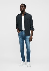 Jeune homme portant une chemise noire ouverte sur un débardeur blanc, un jean slim bleu et des baskets blanches, debout avec une main dans la poche.