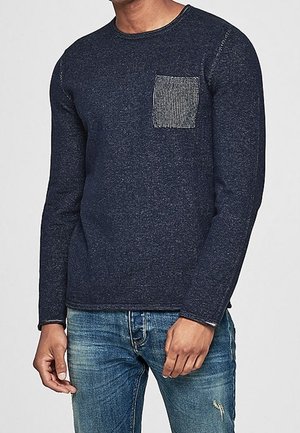 Mann trägt einen marineblauen Pullover mit langen Ärmeln, einer strukturierten grauen Brusttasche und ausgewaschenen blauen Jeans mit einem kleinen Riss nahe am Knie.