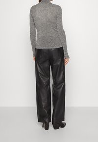 Femme portant un pull gris à col roulé à manches longues, un pantalon large en cuir noir et des bottes à talons noires, debout face à un mur blanc uni.