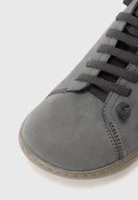 Punta di scarpa in suede grigio con lacci intrecciati grigio scuro e cuciture visibili su suola in gomma grigia chiara, il tutto su uno sfondo bianco.