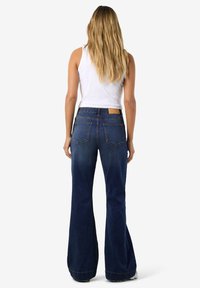 Tamsiai mėlyni platėjančių kojų džinsai su aukštu liemeniu, turintys galines kišenes ir smėlio spalvos odinį prekės ženklo lopelį ant juosmens.