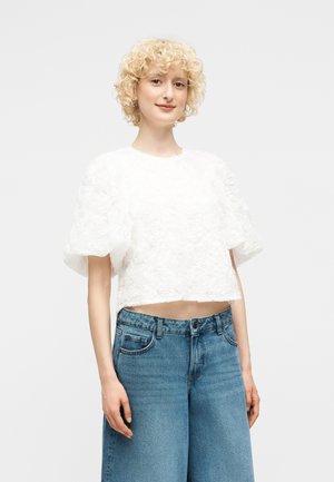 Mladá žena s kučeravými blond vlasmi oblečená v bielom textúrovanom tričku s krátkym rukávom a v modrých rifliach s vysokým pásom stojaca pred jednoduchým pozadím.