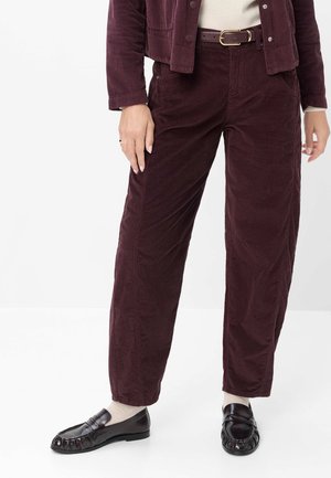 Person iført burgundy kordbuksbukser og jakke, beige skjorte, sorte loafers og beige sokker, stående mod en hvid baggrund.