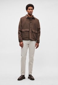 Pruun villa segust jakk kraega, millel on kaks eesmist taskut ja nööpidega kinnis. Sobitatud heledate beežide pükste ja tumedate mokassiinidega.