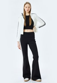 Cropped witte zip hoodie met zwarte strepen, gecombineerd met zwarte flared broek. Model draagt een aangesloten zwart topje en witte sneakers.
