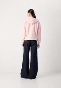 Sweat à capuche rose clair avec un ourlet côtelé associé à un jean large bleu foncé. Le matériau semble doux, avec des détails visibles minimes.