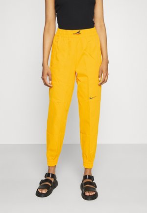 Personne portant un pantalon de jogging Nike jaune vif, un haut noir sans manches et des sandales noires avec boucles, debout sur un sol gris.