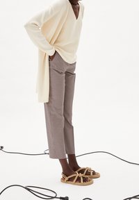 Kermanvärinen neulepusero V-kaula-aukkolla, ruskeat ruudulliset housut ja ruskeat köysipidikkeiset sandaalit. Taustalla mustia johtoja valkoisella pinnalla.